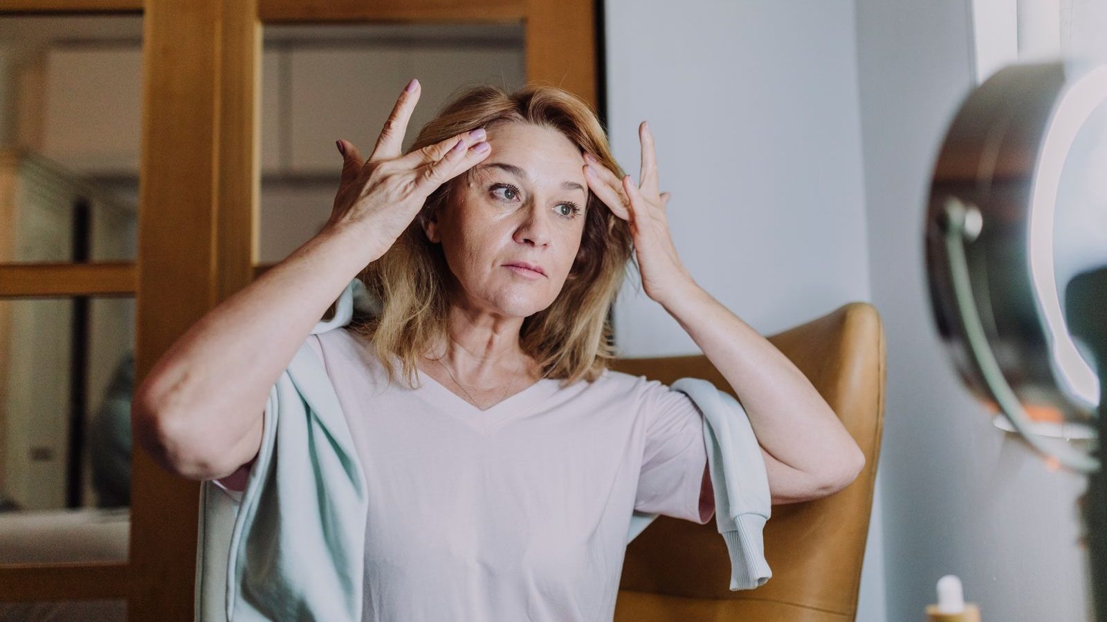 Une femme aux cheveux blonds mi-longs, assise dans un fauteuil, masse ses tempes du bout des doigts devant un miroir lumineux
