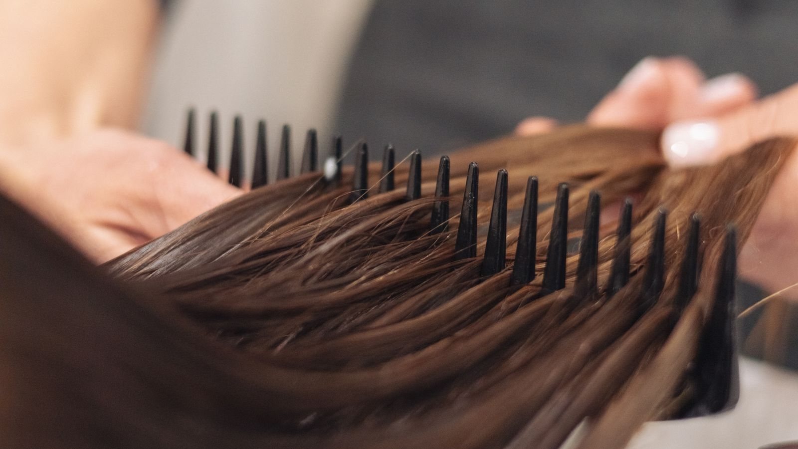 Un peigne à larges dents démêle une mèche de cheveux châtains mouillés, tenue par la main d’une coiffeuse