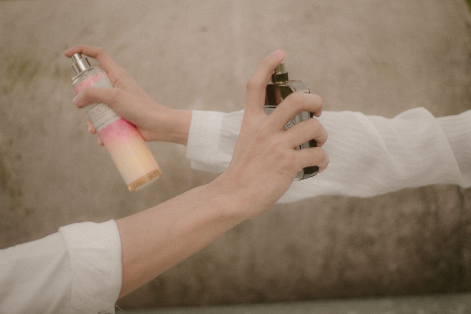 Deux mains vaporisent l’une une brume parfumée en flacon rose, l’autre un parfum en flacon de verre, sur des bras vêtus de blanc