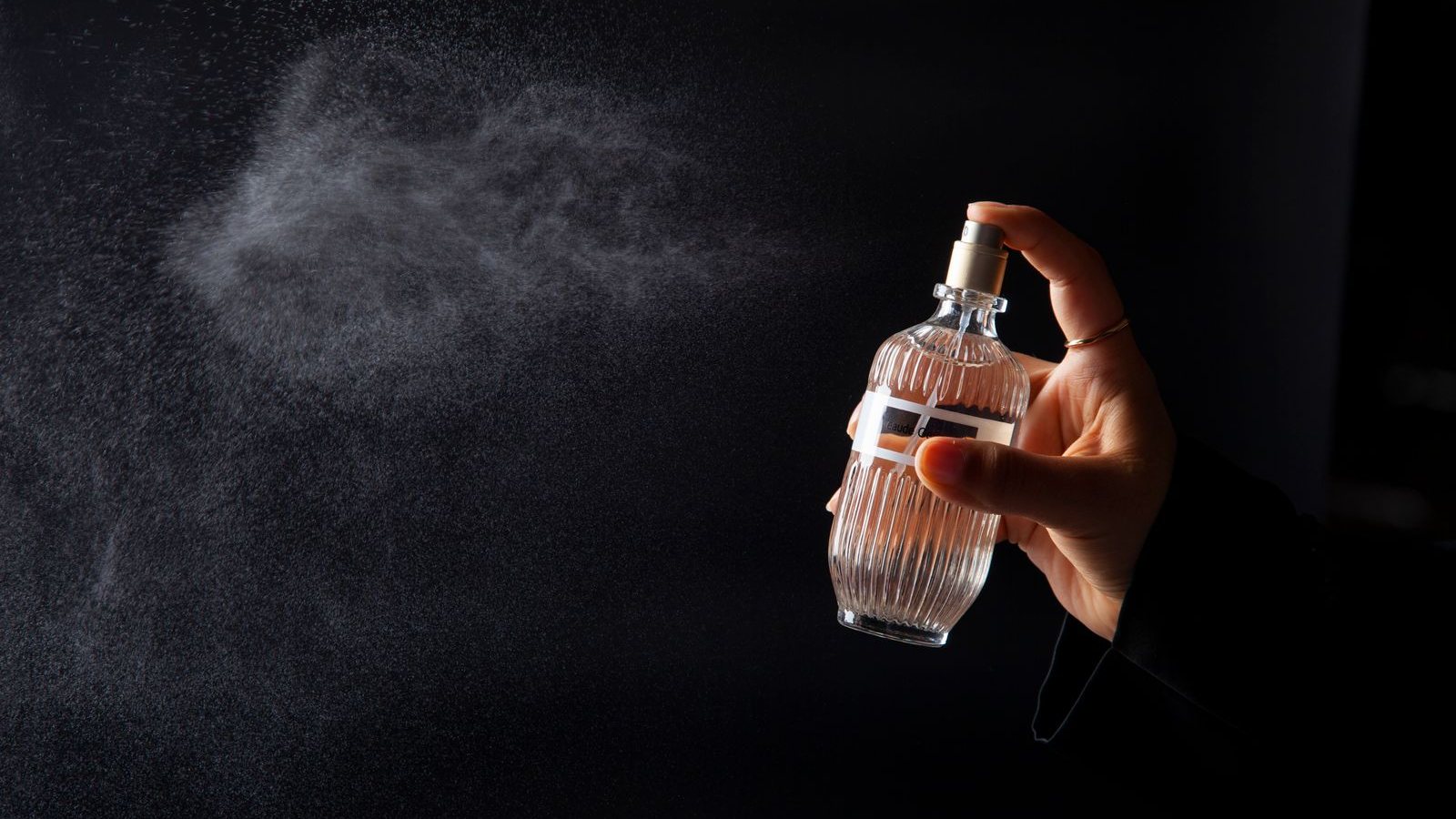 Une main vaporise un flacon de verre strié, dont la fine brume se disperse en nuage sur un fond noir