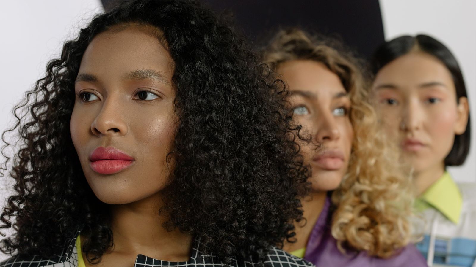 Trois femmes de profil aux cheveux bouclés noirs, bouclés blonds et lisses, alignées l’une derrière l’autre devant un fond clair
