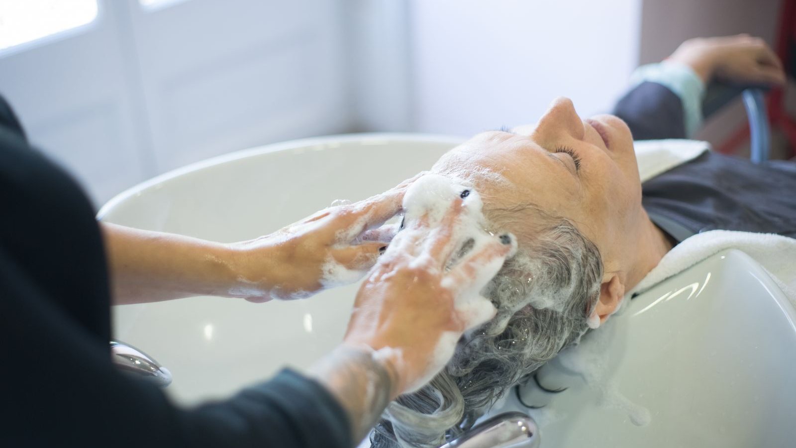 Une coiffeuse masse le cuir chevelu couvert de mousse d’une cliente aux cheveux gris, allongée au bac de lavage d’un salon