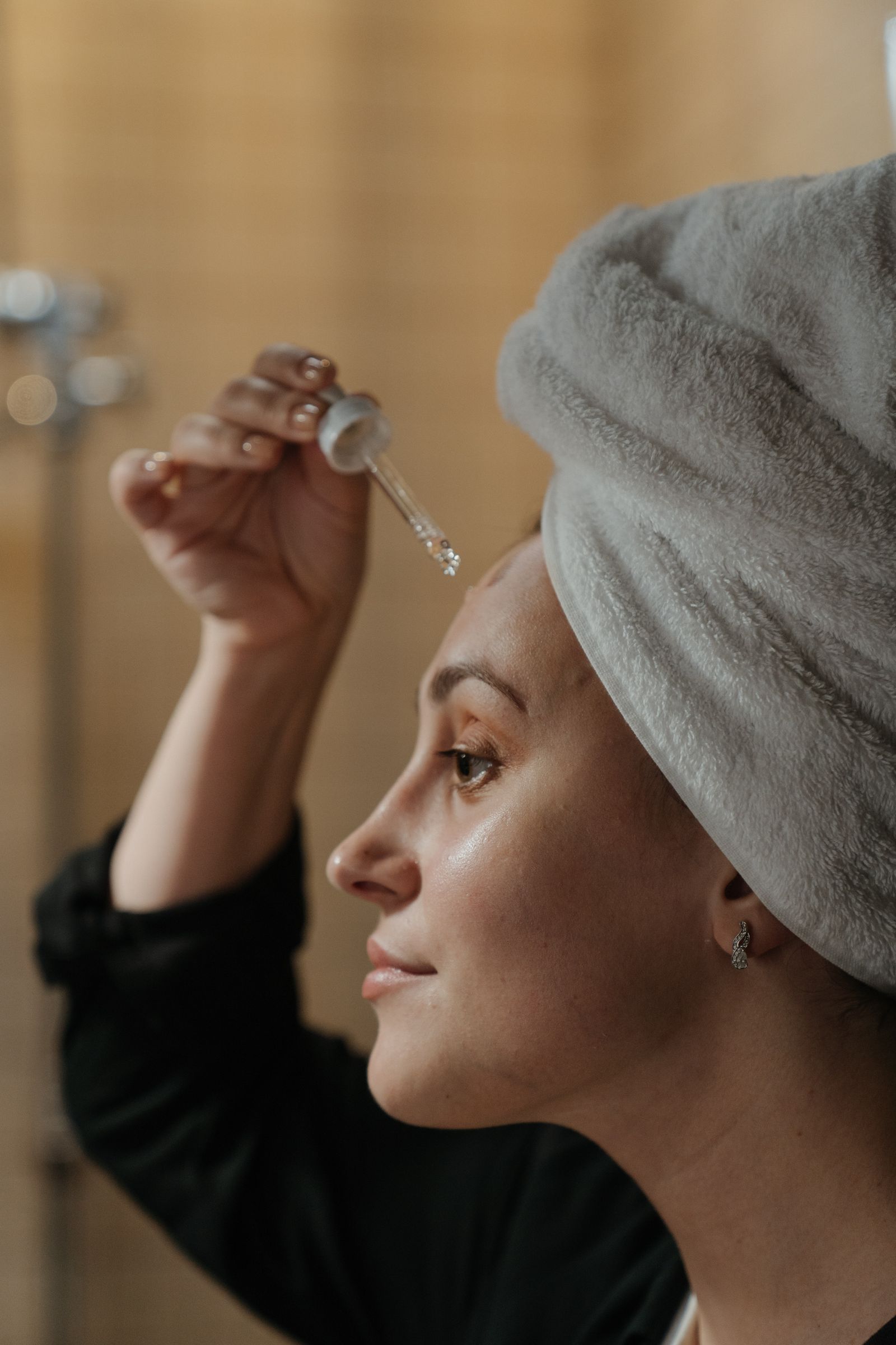 Une personne, serviette sur les cheveux, applique un sérum au compte-gouttes sur son front après le nettoyage