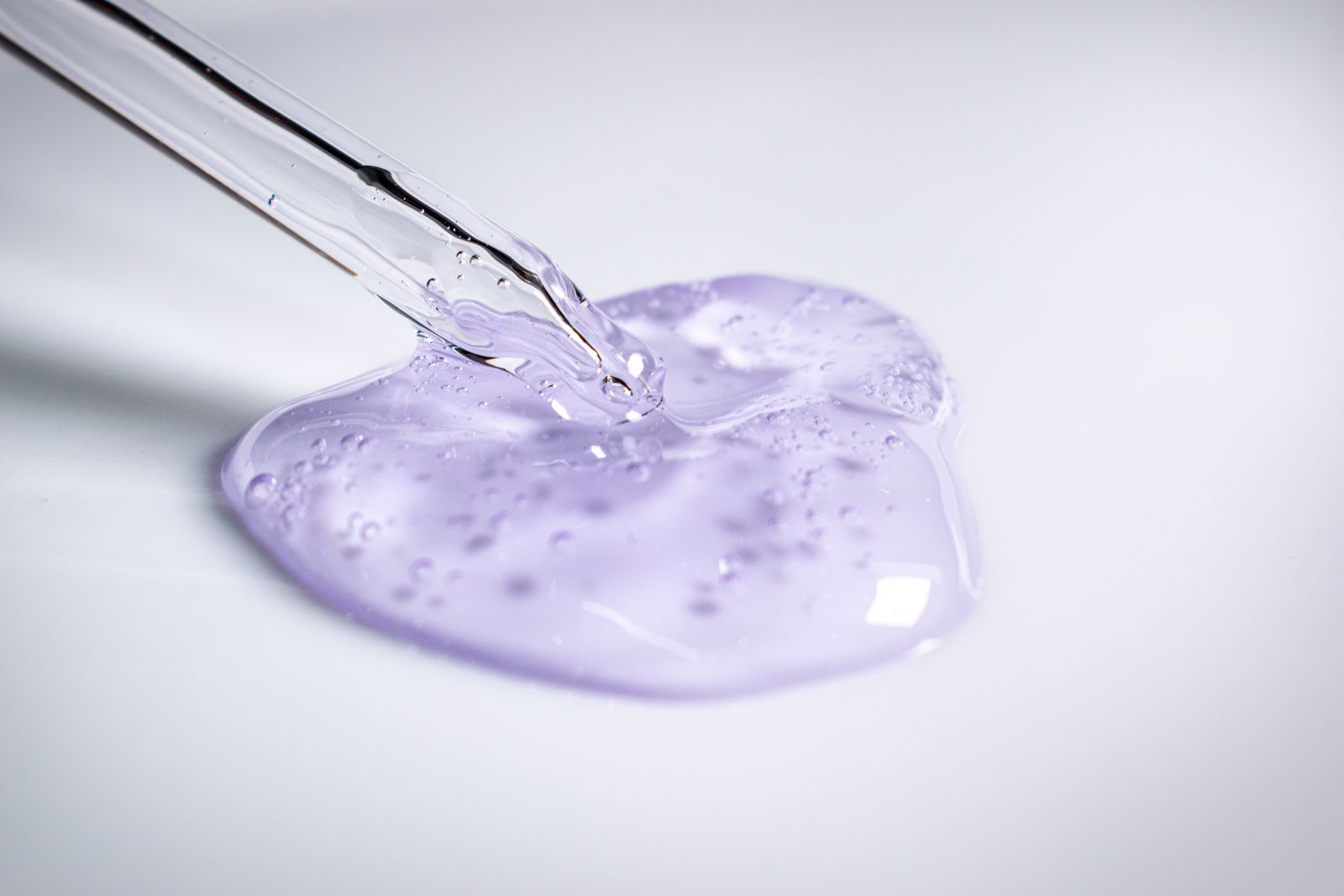 Compte-gouttes en verre déposant une nappe de gel translucide lilas parcourue de bulles, sur fond clair
