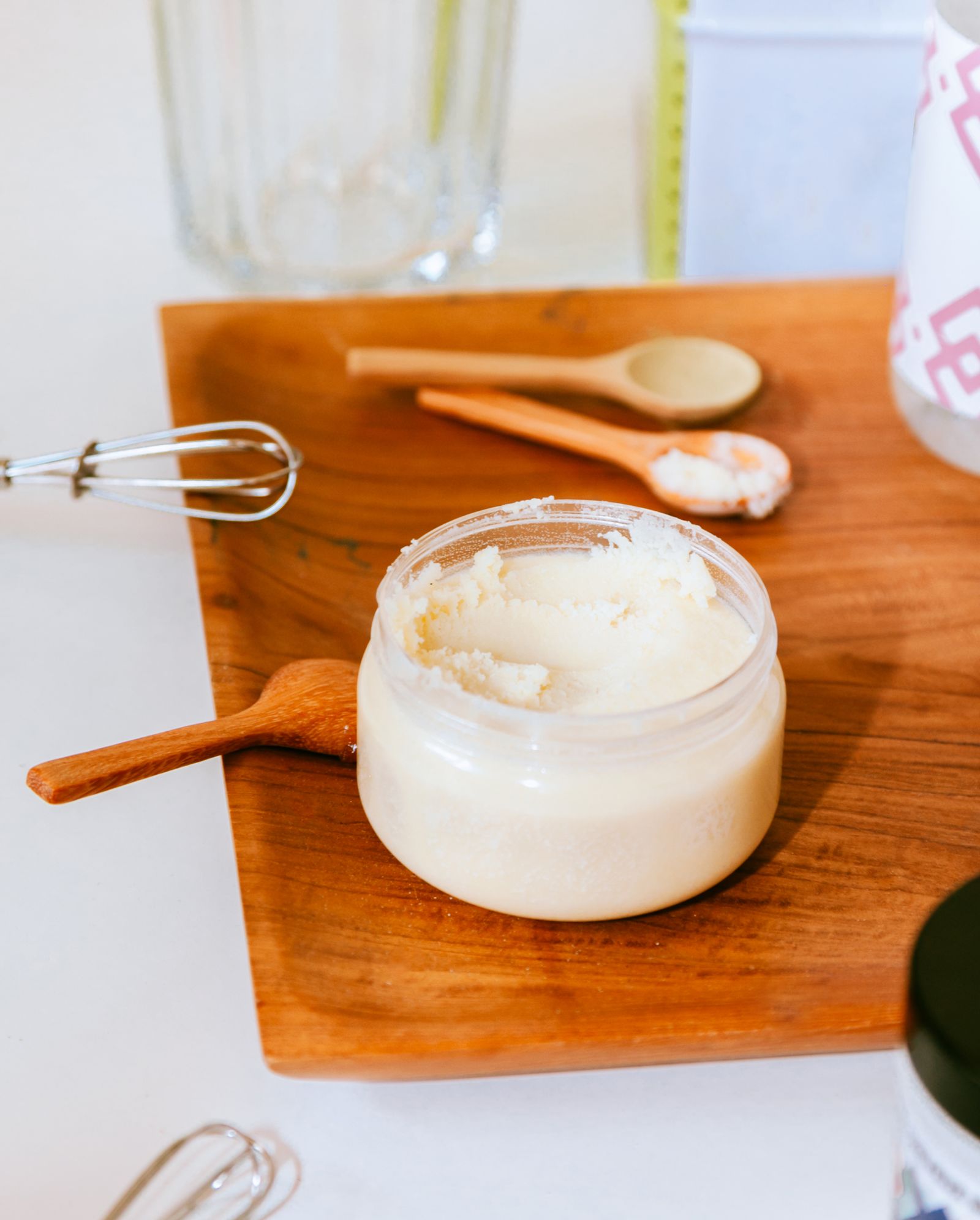 Pot en verre rempli de beurre ivoire à la texture granuleuse, posé sur une planche en bois avec des cuillères et un fouet