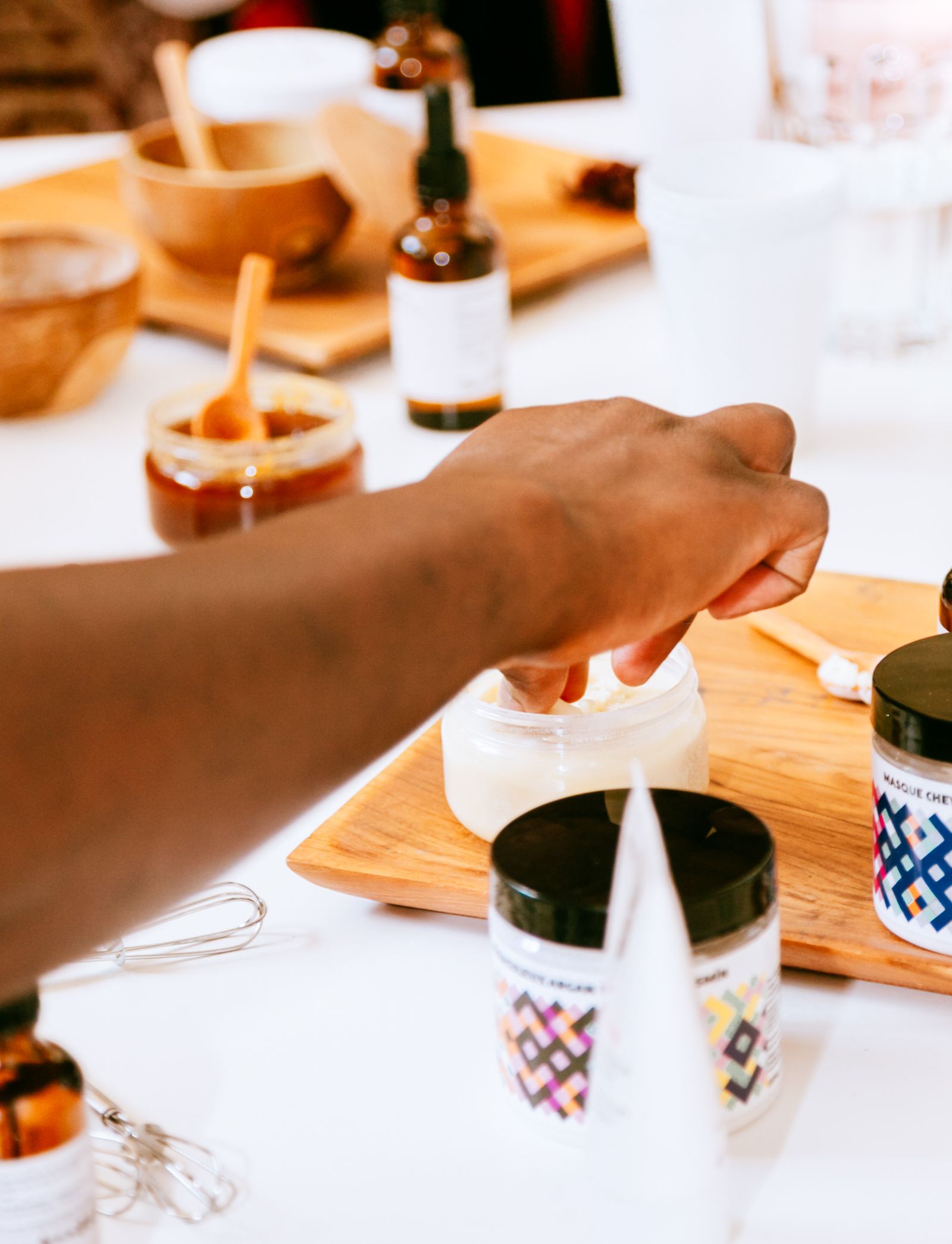Une main prélève du beurre ivoire dans un pot en verre posé sur une planche en bois, entouré d’autres pots de soin