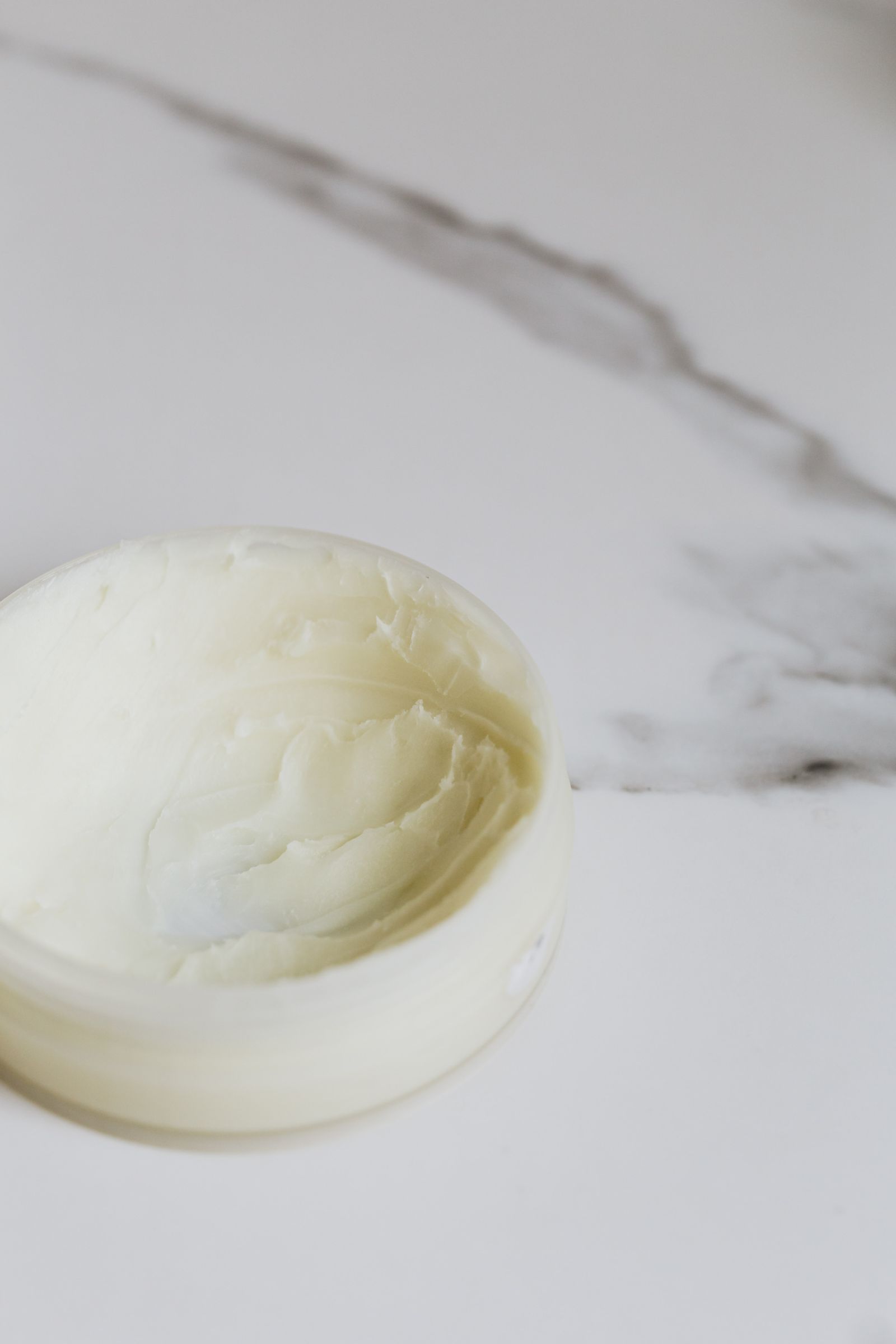Pot ouvert vu de dessus, rempli d’un beurre épais de couleur ivoire tirant sur le jaune pâle, posé sur du marbre blanc