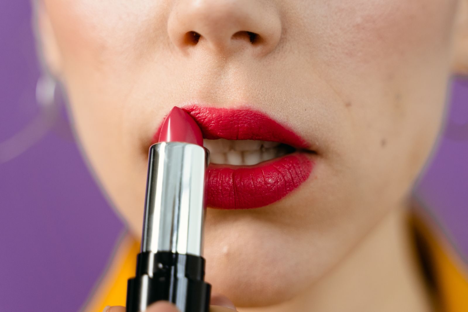 Gros plan sur une bouche pendant l’application d’un rouge à lèvres framboise en bâton, devant un fond violet
