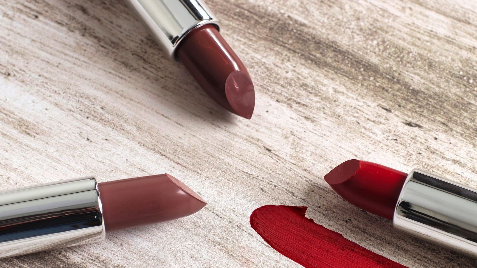 Trois rouges à lèvres en bâton, un brun rosé, un nude et un rouge vif, posés sur un plan clair à côté d’un trait de rouge
