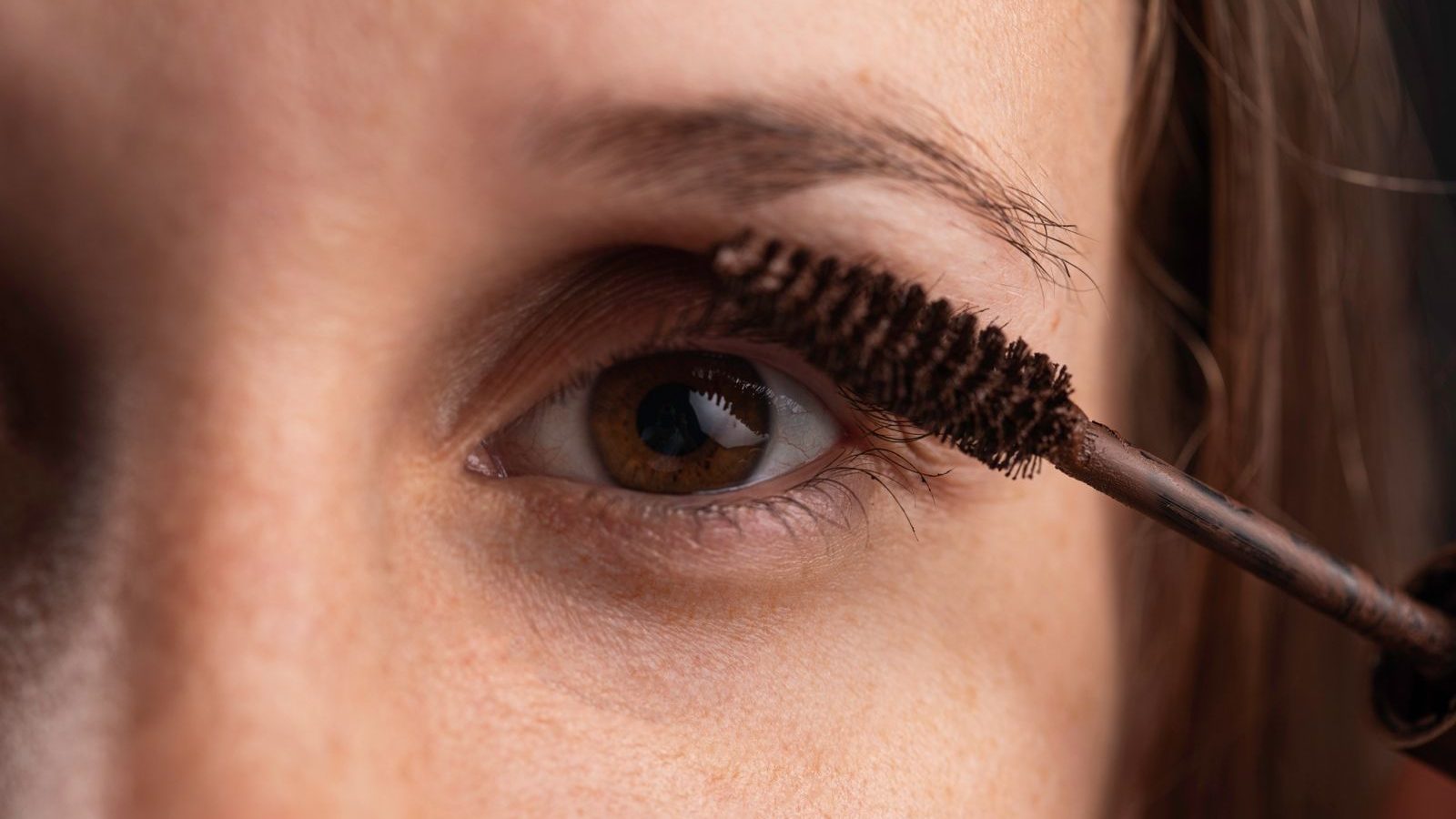 Gros plan sur un œil aux iris marron pendant qu’une brosse de mascara brun gaine les cils du haut