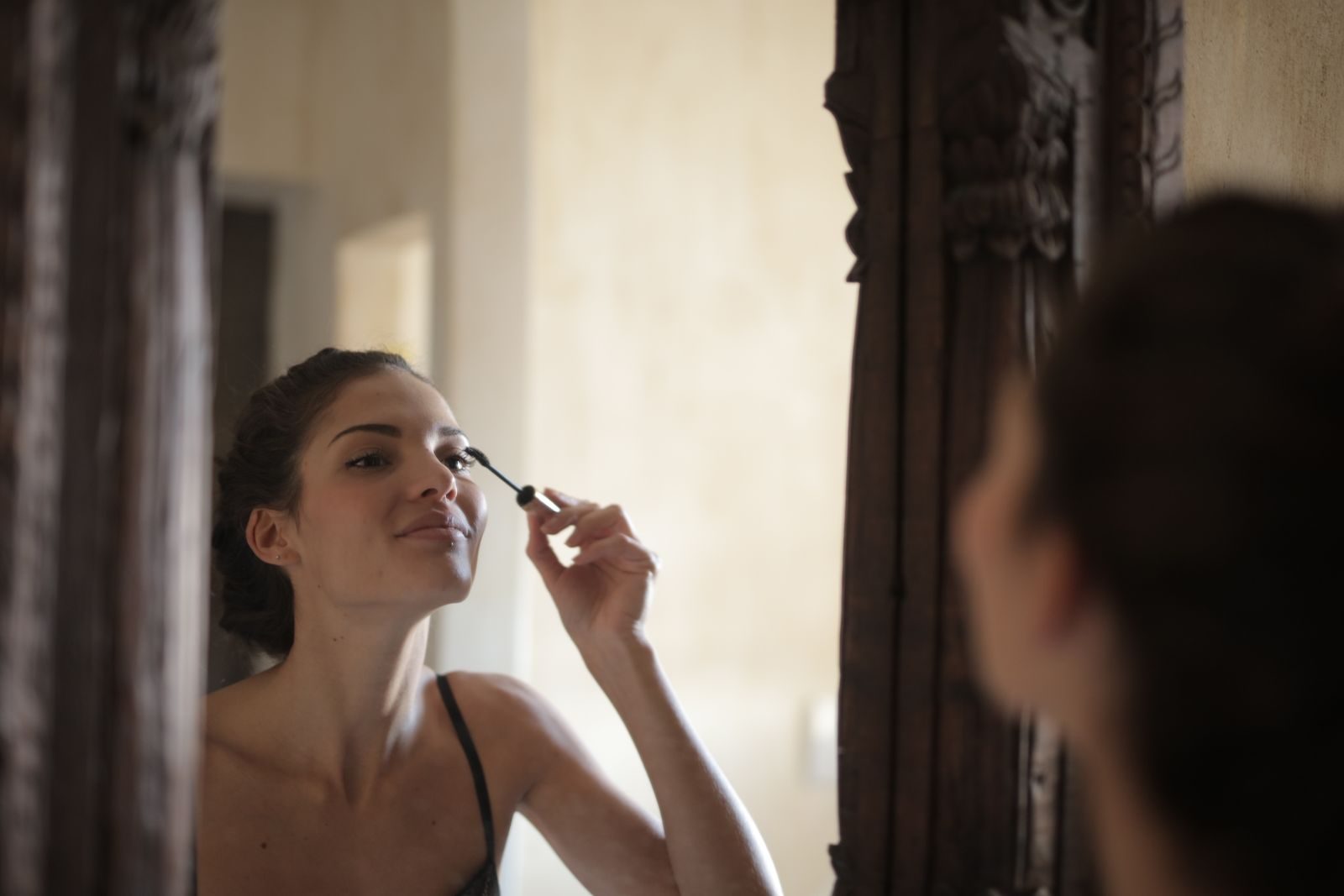 Face à un miroir au cadre de bois sculpté, une jeune femme souriante applique du mascara sur ses cils