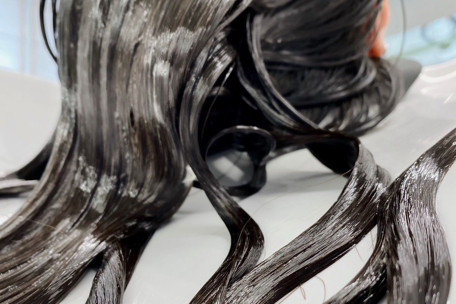 De longues mèches de cheveux bruns enduites de masque capillaire, étalées au bac de lavage d’un salon
