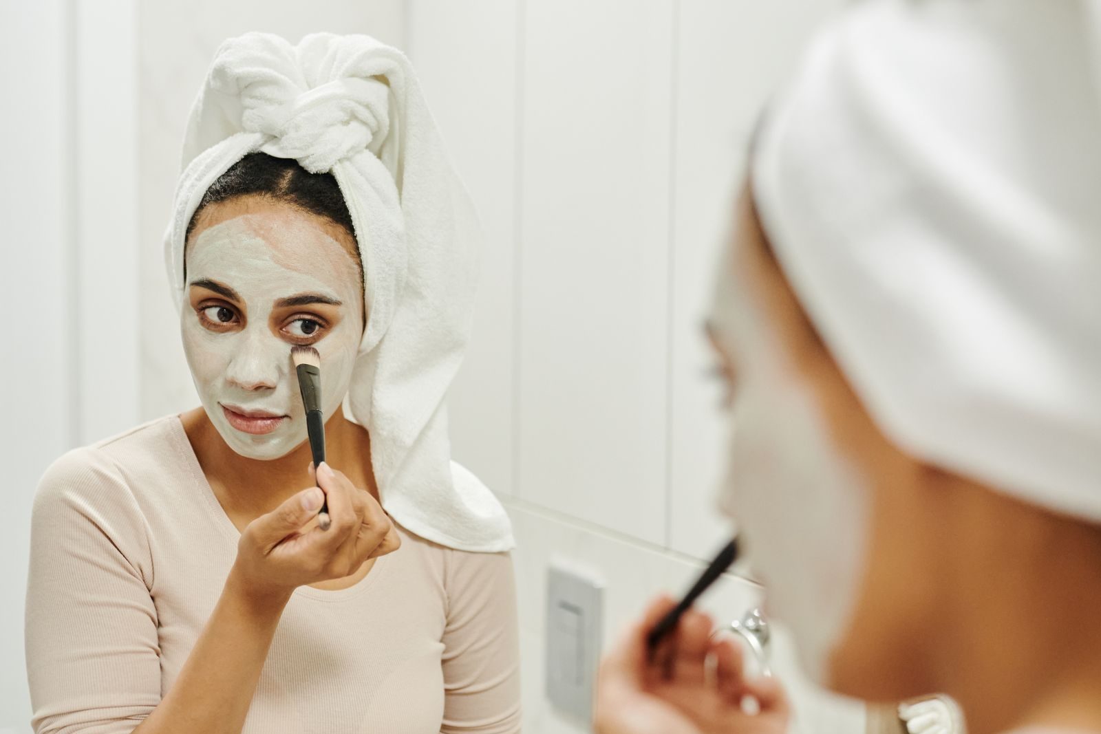 Face au miroir de sa salle de bains, une femme aux cheveux enveloppés dans une serviette étale au pinceau un masque de couleur claire sur son visage