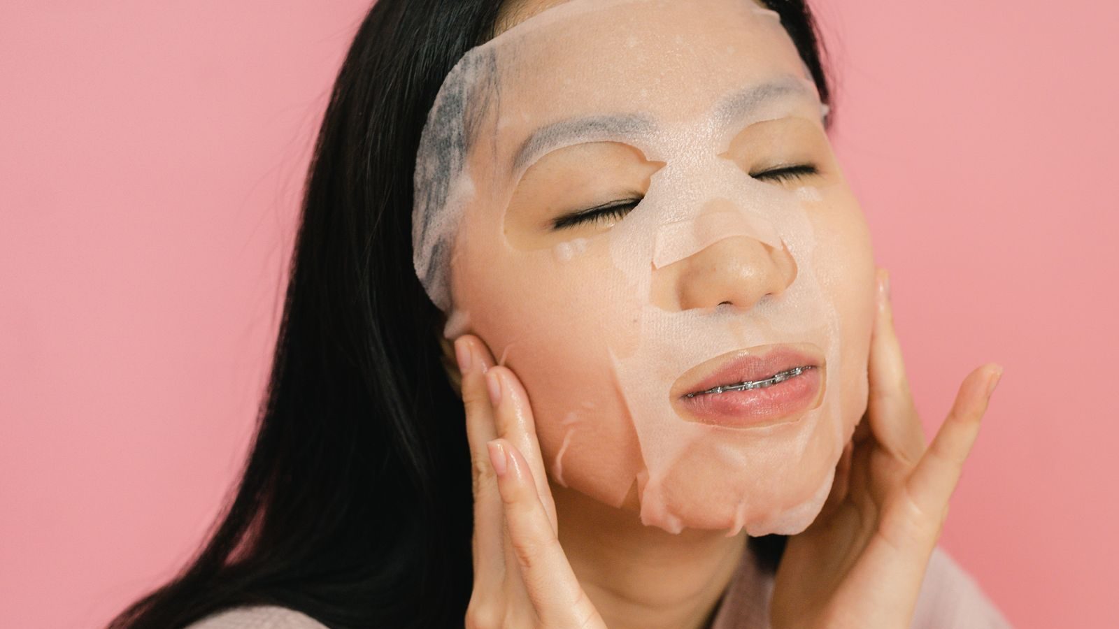 Une jeune femme aux yeux fermés lisse du bout des doigts un masque en tissu posé sur son visage, devant un fond rose