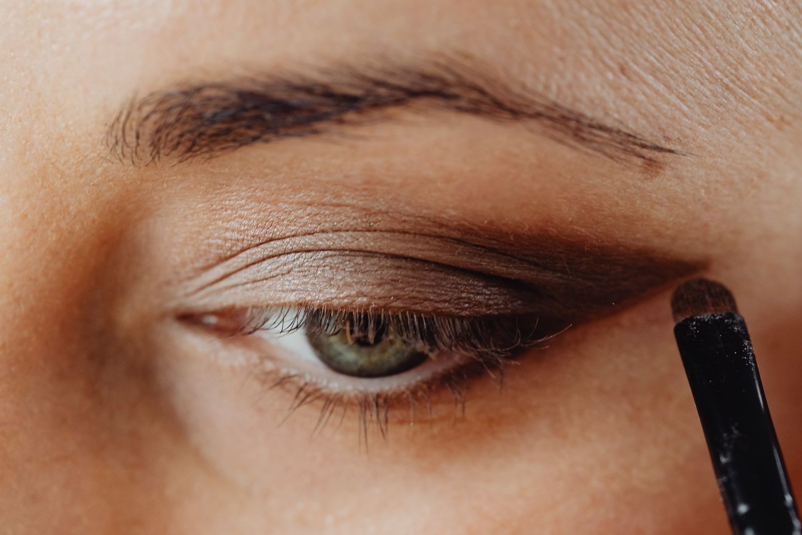 Gros plan sur un œil vert dont la paupière reçoit un fard brun mat, estompé vers l’extérieur avec un petit pinceau
