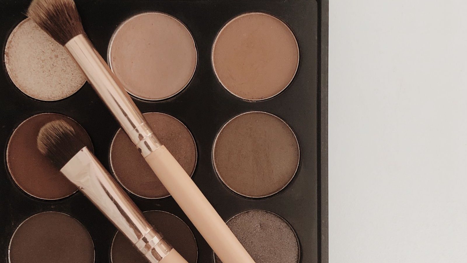 Palette de fards à paupières aux teintes beige, taupe et brun, mates et irisées, avec deux pinceaux posés en travers des godets