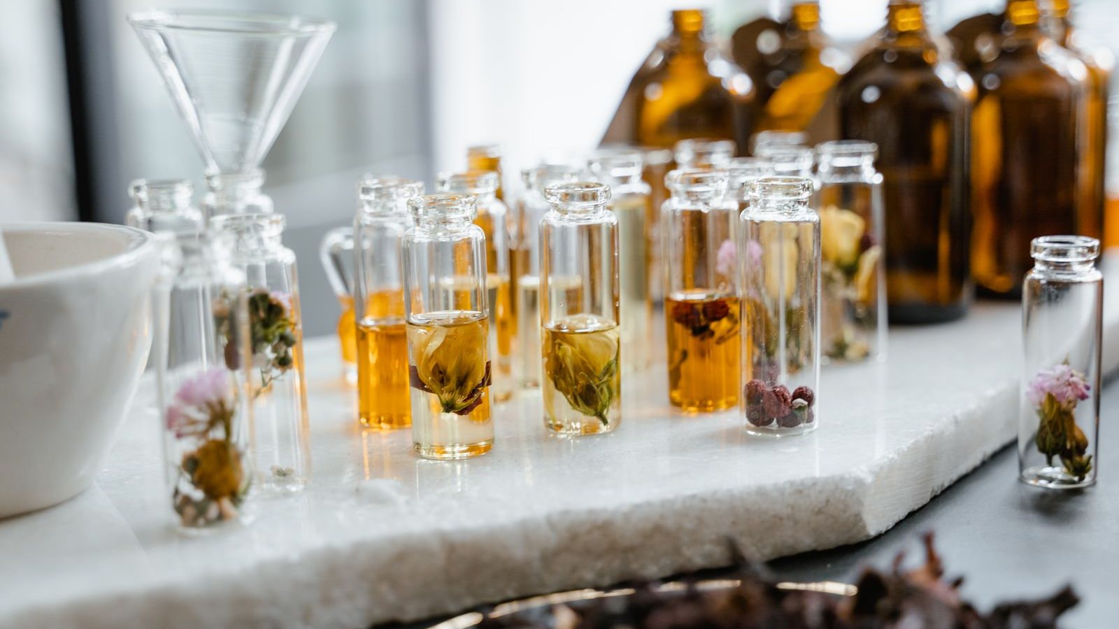Des fioles de verre remplies de jus ambrés et de fleurs séchées, alignées sur une plaque de marbre devant des flacons bruns de parfumeur