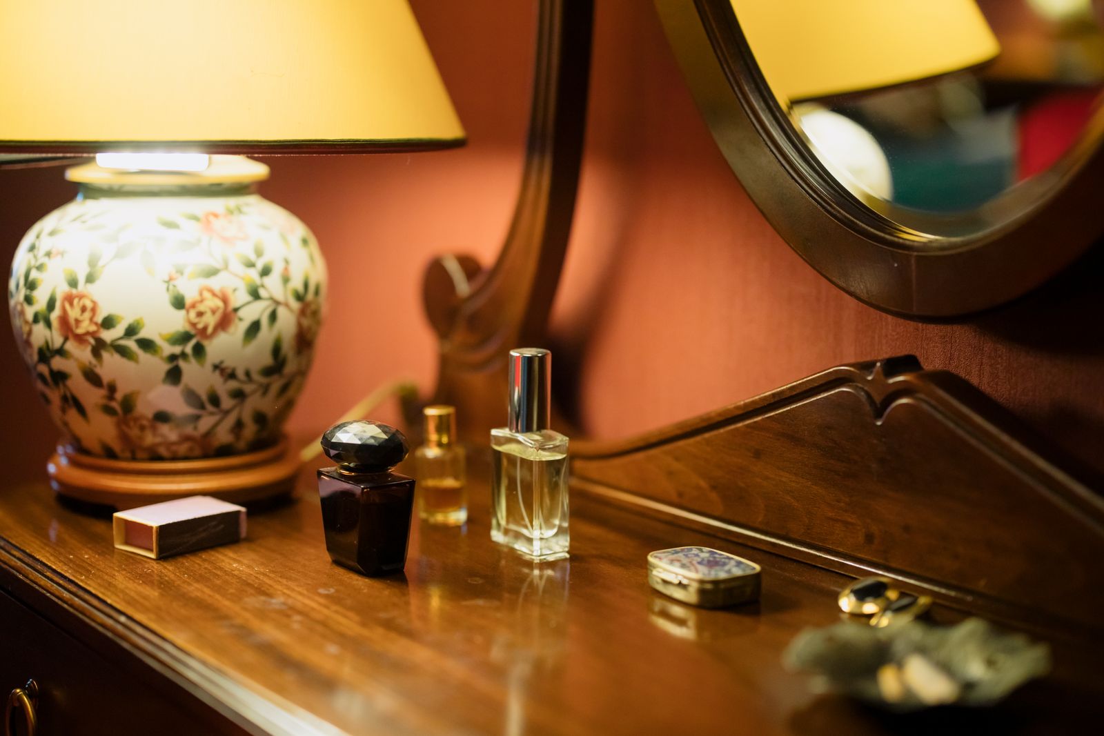 Plusieurs petits flacons de parfum posés sur une coiffeuse en bois, devant un miroir, éclairés par une lampe de chevet
