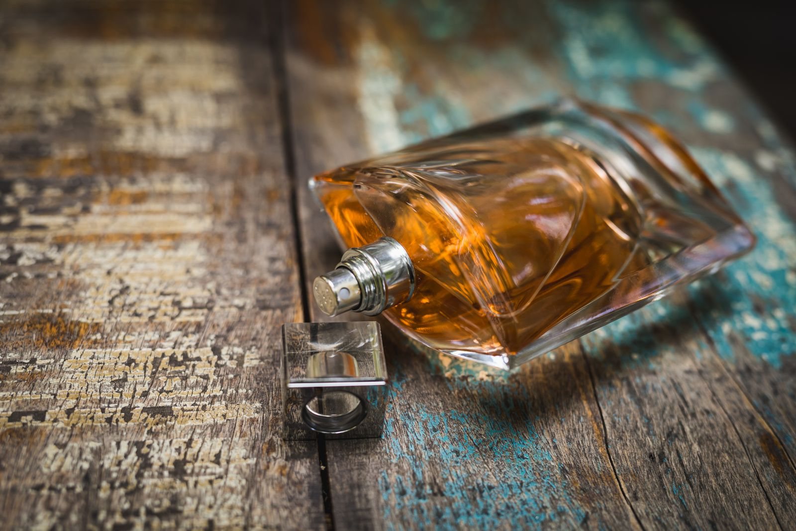 Un flacon de parfum au jus ambré couché sur une table en bois patiné, à côté de son bouchon métallique carré