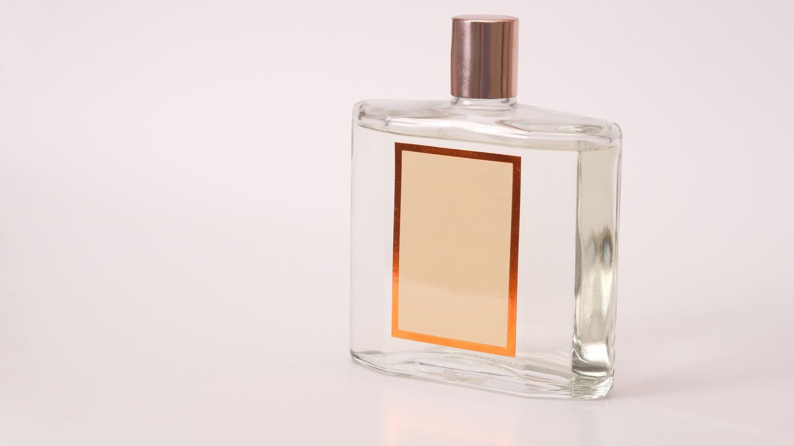 Un flacon de parfum rectangulaire au bouchon cuivré et à l’étiquette vierge, posé sur un fond rose pâle