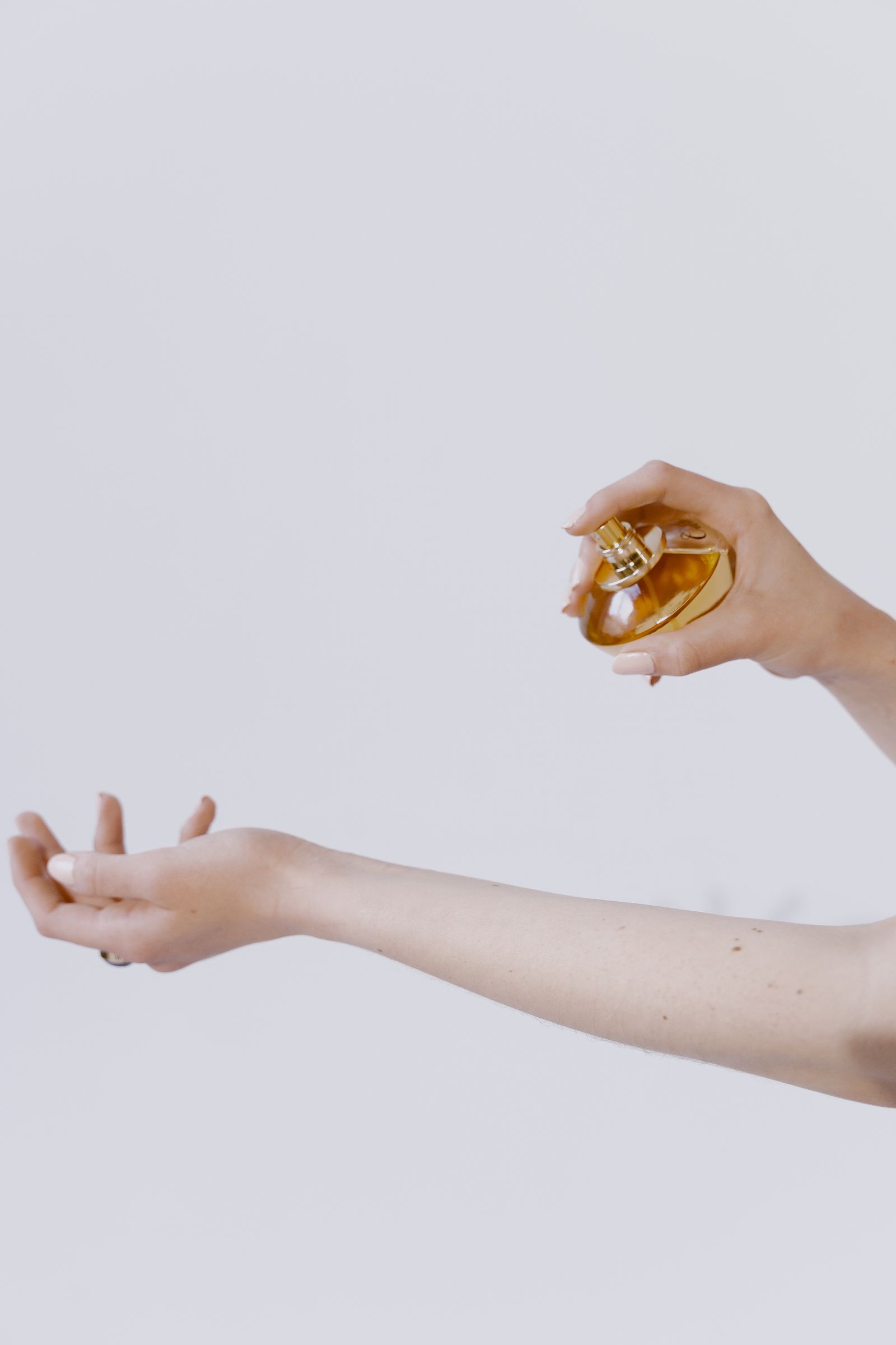 Une main vaporise un flacon de parfum doré sur l’intérieur de l’avant-bras tendu, sur fond blanc