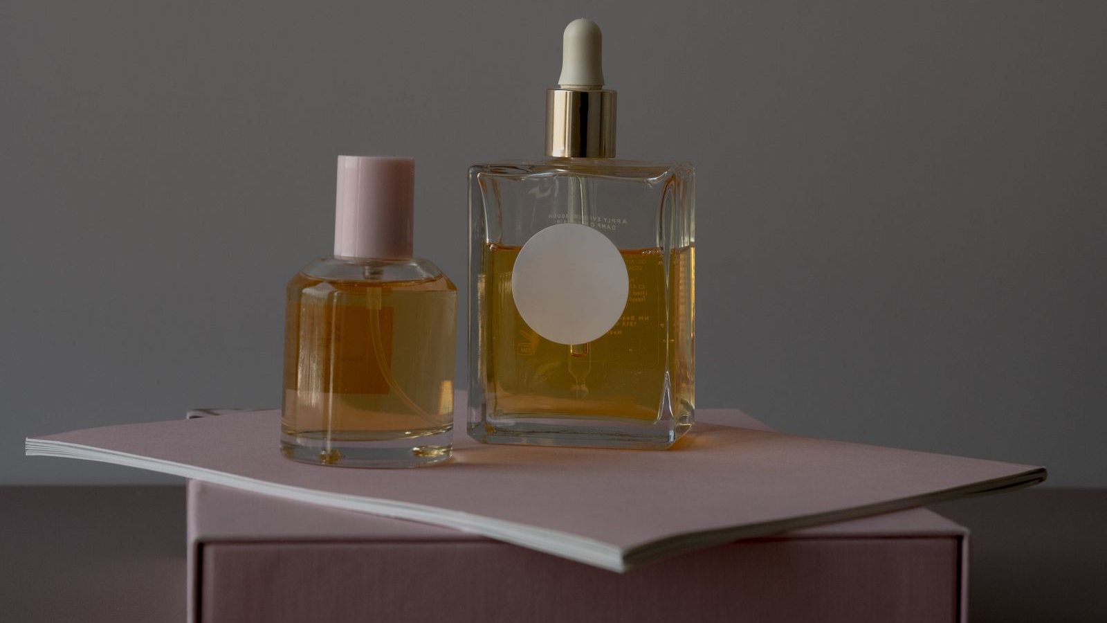 Un flacon de parfum au bouchon rose et un flacon à pipette rempli d’un jus ambré, posés sur un carnet rose au-dessus d’une boîte
