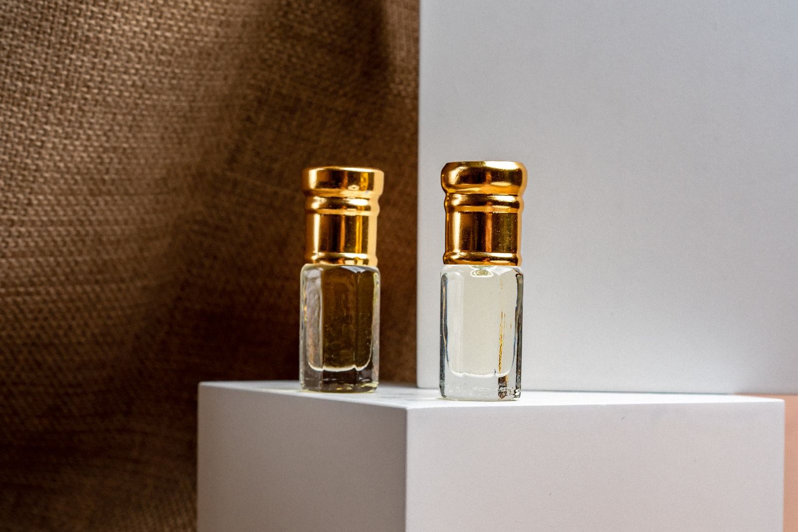 Deux petits flacons de parfum à l’huile, au bouchon doré, posés sur un cube blanc devant une toile de jute