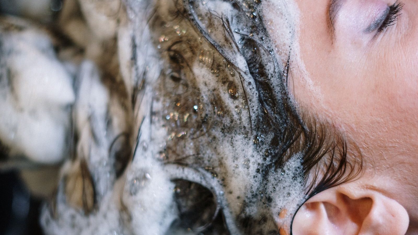 Une femme aux yeux fermés, les cheveux couverts de mousse de shampoing, allongée au bac de lavage d’un salon