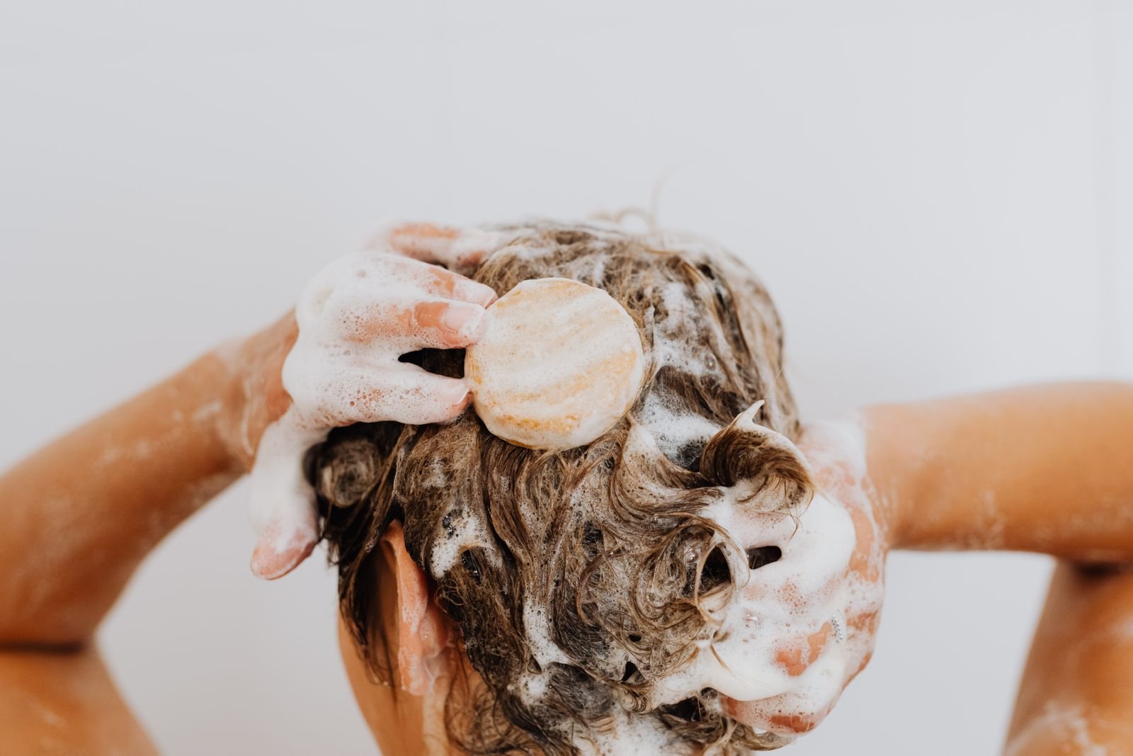 Une personne savonne ses cheveux mouillés avec un pain de shampoing solide rond, la tête couverte de mousse blanche