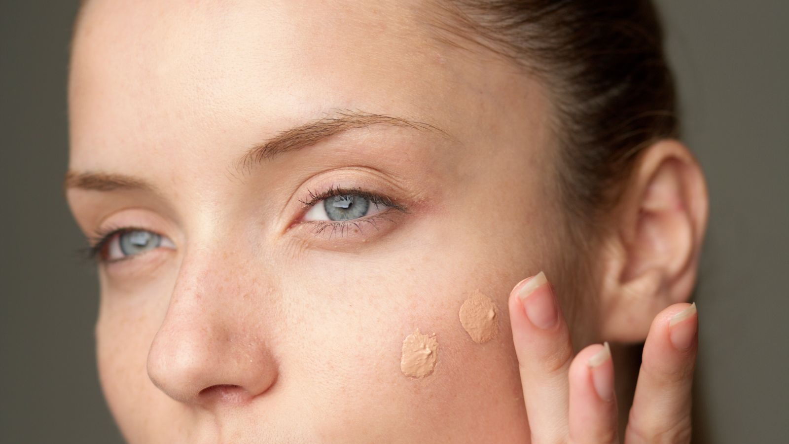 Gros plan sur le visage d’une femme, deux touches de fond de teint déposées sur la joue, la main levée près de la tempe