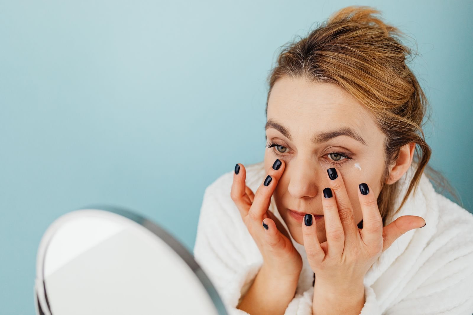 Une femme en peignoir blanc tapote une crème sous ses yeux du bout des doigts, face à un miroir rond