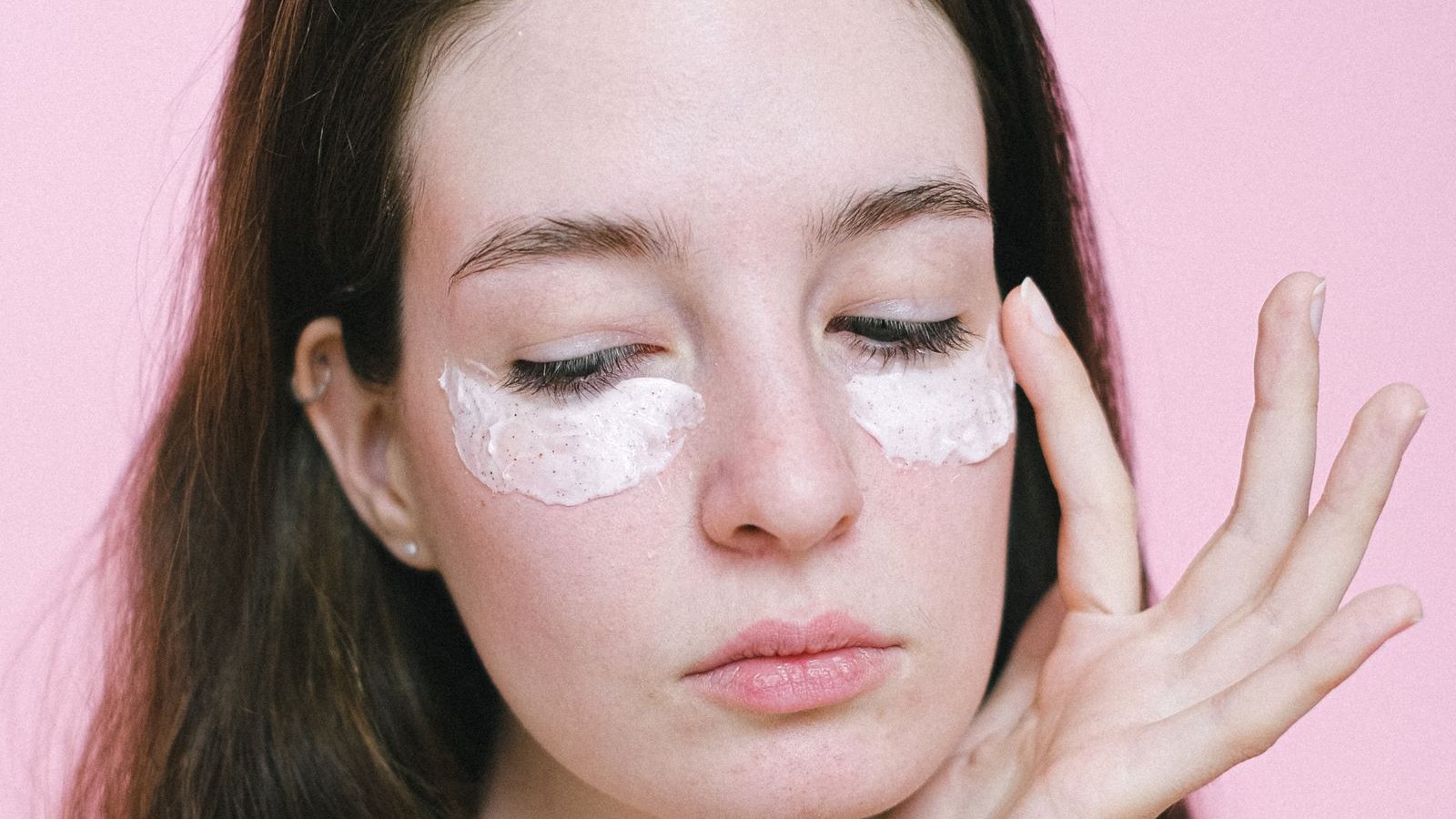 Une jeune femme aux yeux fermés, une couche de crème claire sous chaque œil, effleure sa tempe du bout des doigts